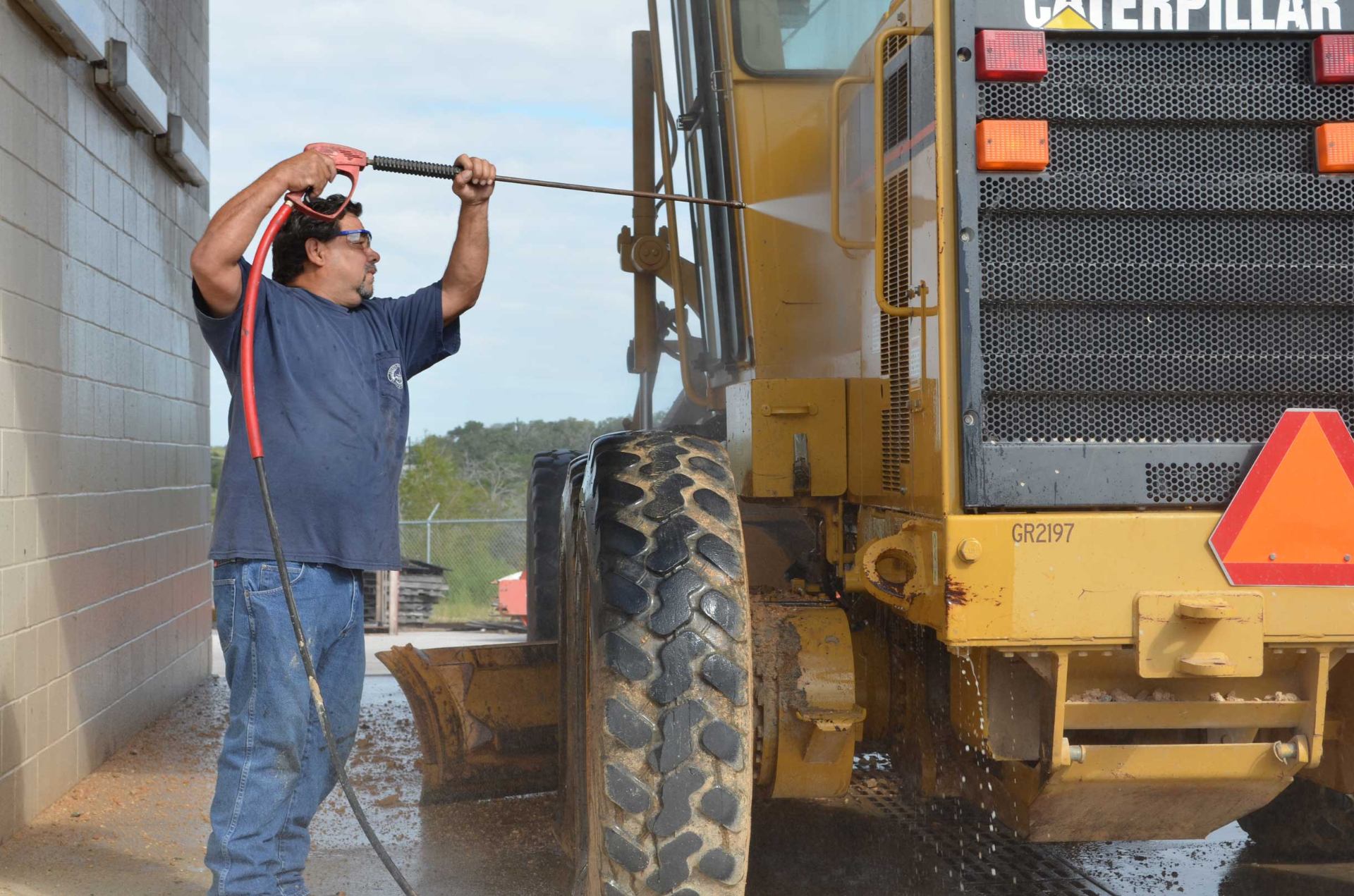 Operator using an Enzo's Cleaning Solutions hot water pressure washer to clean a Caterpillar motor grader in a construction equipment wash bay